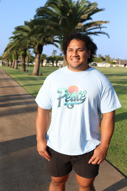 Person wearing a light blue t-shirt with a graphic design, standing outdoors with palm trees in the background.