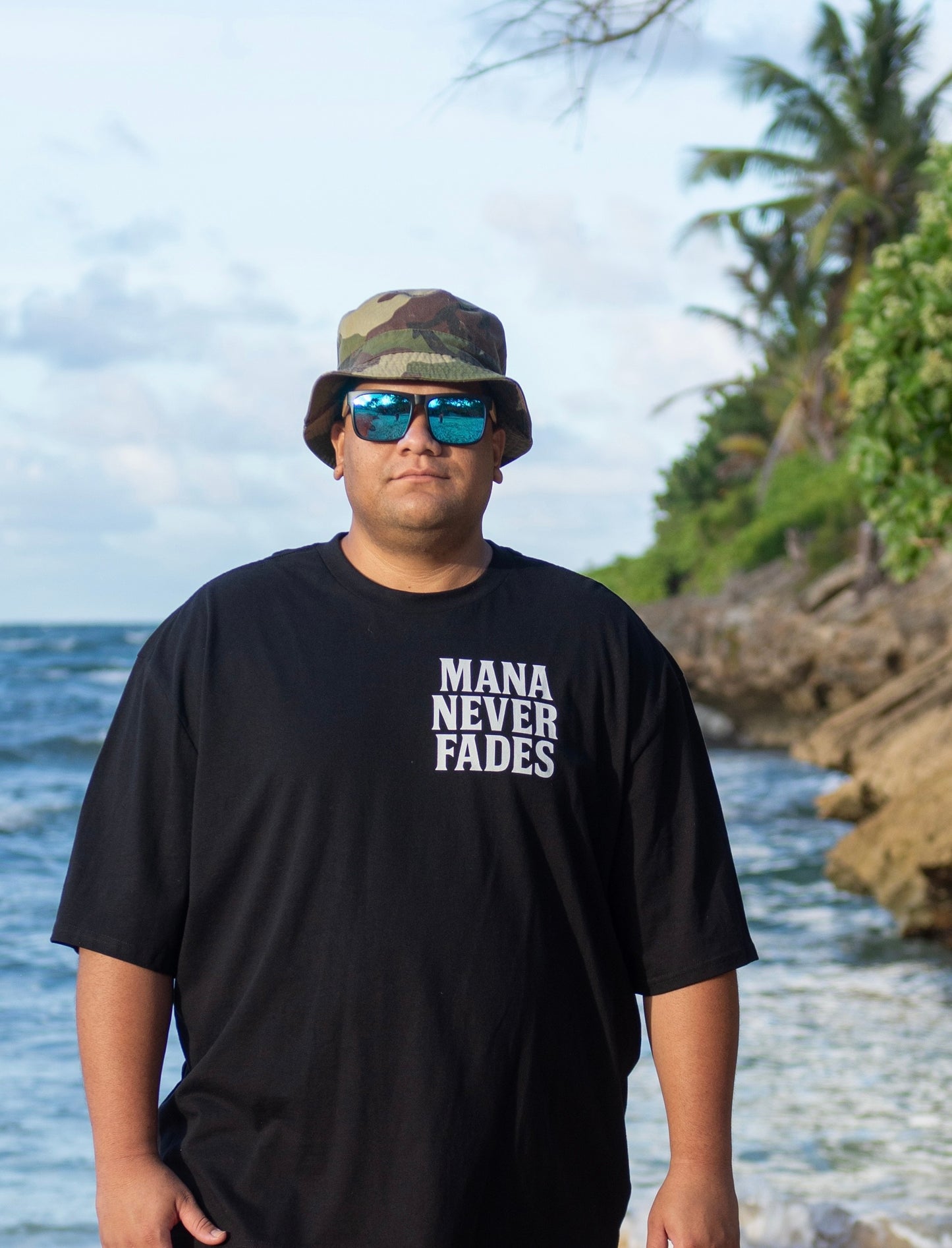 Person wearing a black t-shirt with 'Mana Never Fades' text, sunglasses, and a camouflage hat standing by a body of water.