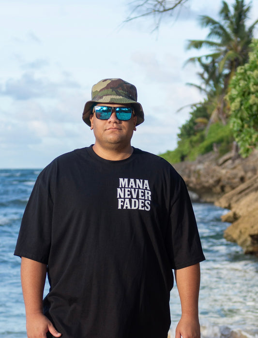 Person wearing a black t-shirt with 'Mana Never Fades' text, sunglasses, and a camouflage hat standing by a body of water.